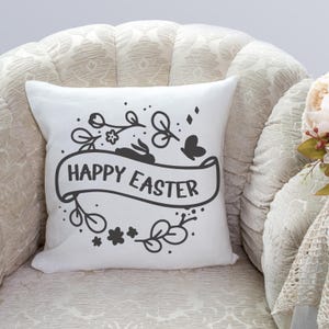 May include: White cushion with a black and white design featuring a bunny rabbit, flowers, and a banner that reads "Happy Easter".