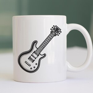 May include: White ceramic mug featuring a black and white illustration of an electric guitar. The guitar design is detailed, showing the strings, pickups, and tuning pegs. The mug has a curved handle and a smooth, glossy finish.