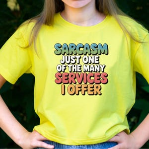 May include: A yellow t-shirt with the text "SARCASM JUST ONE OF THE MANY SERVICES I OFFER" in a colorful, retro font. The shirt is a bright, solid color and has short sleeves.