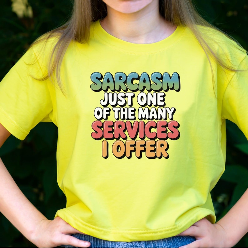 May include: A yellow t-shirt with the text "SARCASM JUST ONE OF THE MANY SERVICES I OFFER" in a colorful, retro font. The shirt is a bright, solid color and has short sleeves.