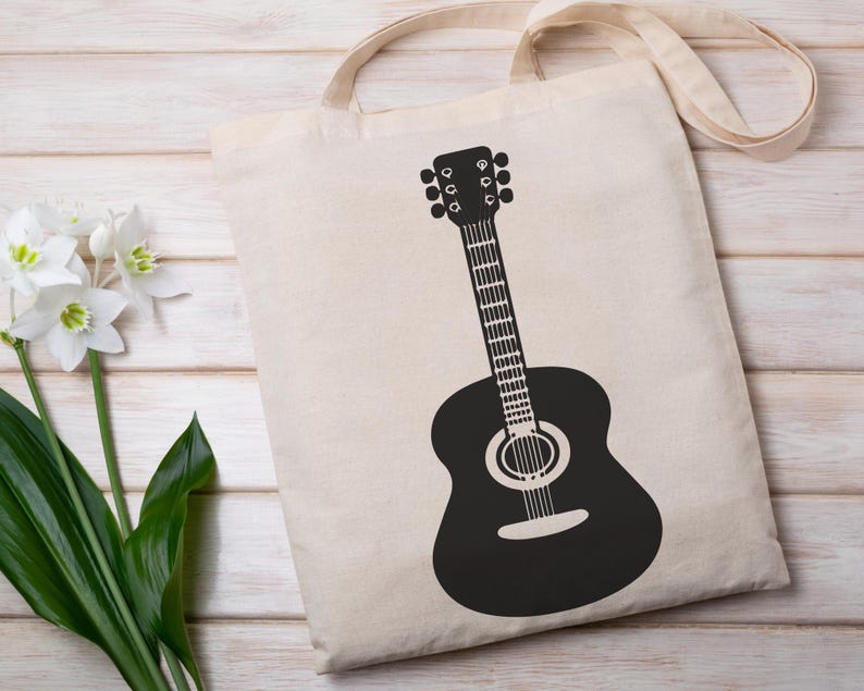 May include: A beige tote bag with a black guitar graphic. The guitar is a detailed silhouette, featuring the body, neck, and headstock. The bag has a simple design with a handle for carrying. The background is a light wooden surface.
