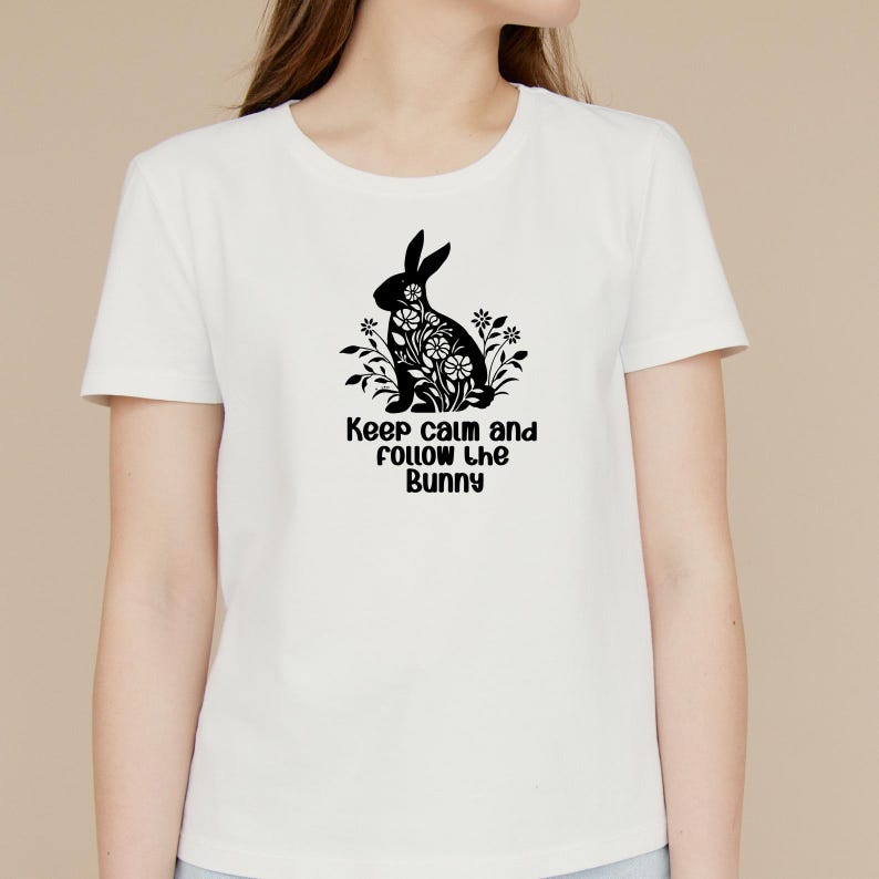 May include: White t-shirt featuring a black silhouette of a bunny rabbit with flowers inside its body. The text "Keep calm and follow the bunny" is printed below the bunny.