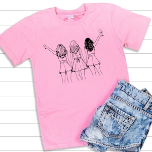 May include: Pink t-shirt with a black line drawing of three women standing together. The woman on the right is holding a glass of wine.