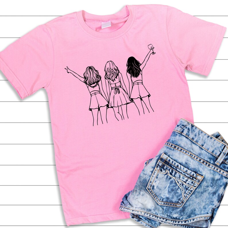 May include: Pink t-shirt with a black line drawing of three women standing together. The woman on the right is holding a glass of wine.