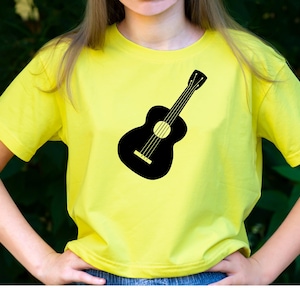 May include: A yellow t-shirt featuring a black guitar silhouette. The guitar design is centered on the shirt. The shirt has short sleeves and a crew neck. The background is a blurred green.