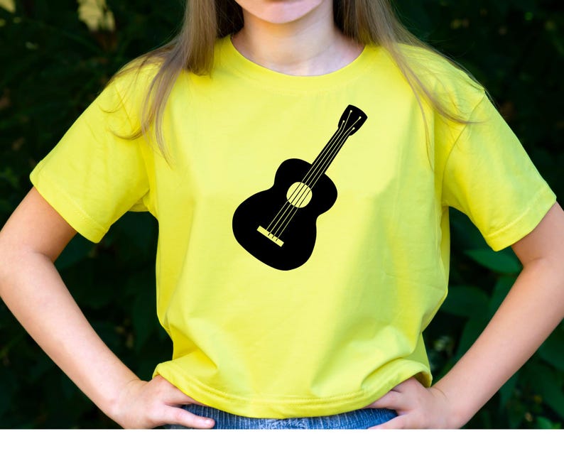 May include: A yellow t-shirt featuring a black guitar silhouette. The guitar design is centered on the shirt. The shirt has short sleeves and a crew neck. The background is a blurred green.