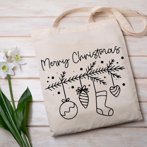 May include: A white canvas tote bag with a black design of a Christmas tree with ornaments and a stocking hanging from the branches. The text "Merry Christmas" is written above the tree.