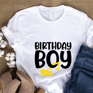 May include: White t-shirt with black text that says "Birthday Boy" and a yellow construction vehicle graphic.