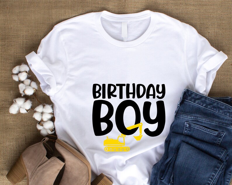 May include: White t-shirt with black text that says "Birthday Boy" and a yellow construction vehicle graphic.