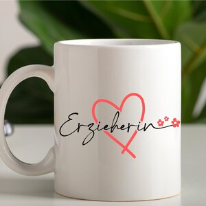 Peut inclure: Une tasse en c&eacute;ramique blanche avec une inscription cursive noire "Erzieherin" et un c&oelig;ur rose avec de petites fleurs roses.