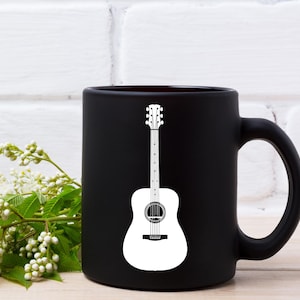May include: A black ceramic coffee mug with a white acoustic guitar graphic. The guitar design is centered on the mug and features a detailed outline. The mug has a curved handle and is set against a white brick wall.