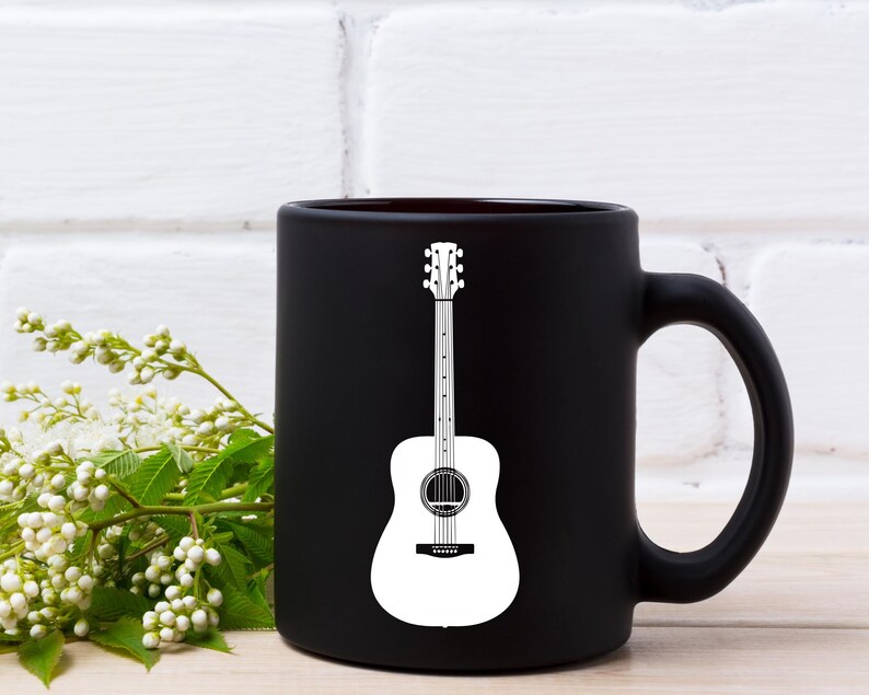 May include: A black ceramic coffee mug with a white acoustic guitar graphic. The guitar design is centered on the mug and features a detailed outline. The mug has a curved handle and is set against a white brick wall.