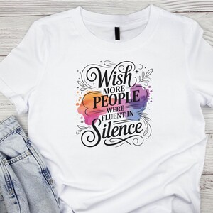 May include: White t-shirt with the text "Wish more people were fluent in Silence" in a decorative font. The text is over a colorful watercolor-style design with orange, pink, purple, and blue hues. The shirt is folded over a pair of blue jeans.