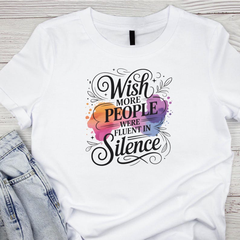 May include: White t-shirt with the text "Wish more people were fluent in Silence" in a decorative font. The text is over a colorful watercolor-style design with orange, pink, purple, and blue hues. The shirt is folded over a pair of blue jeans.