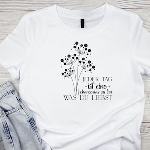 May include: White t-shirt with a black graphic design of three dandelion-like flowers and the text "JEDER TAG ist eine chance das zu tun WAS DU LIEBST".