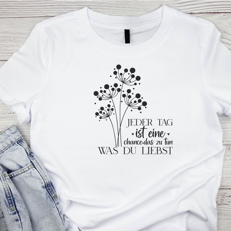 May include: White t-shirt with a black graphic design of three dandelion-like flowers and the text "JEDER TAG ist eine chance das zu tun WAS DU LIEBST".