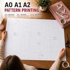 May include: A large white pattern printing sheet with garment outlines, held by hands. Text at the top reads "A0 A1 A2 PATTERN PRINTING". Additional text includes "TRUE SCALE PRINTS TO 100%" and "NO TAPING READY TO CUT & USE". A pink fabric and scissors are also visible.