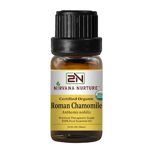 Op de afbeelding: Een kleine amberkleurige glazen fles met essentiële olie van Romeinse kamille van Nirvana Nurture. Het etiket luidt "Certified Organic" en "100% Pure Essential Oil". De fles heeft een zwarte dop en bevat 10 ml olie.