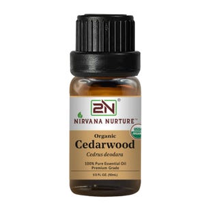 Op de afbeelding: Een kleine amberkleurige glazen fles met biologische essentiële olie van cederhout. Het etiket luidt "Nirvana Nurture" en "Cedarwood". De fles heeft een zwarte dop en bevat 10 ml 100% pure essentiële olie.