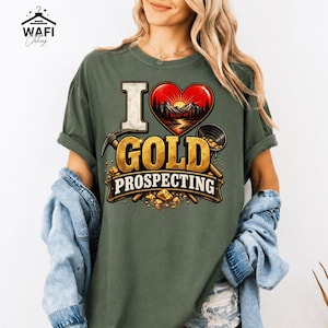 Peut inclure: Un t-shirt vert olive avec un motif imprimé indiquant I LOVE GOLD PROSPECTING. Le design comprend un cœur rouge avec un paysage de montagne, une pioche, une batée et des pépites d'or. Le texte est écrit dans une police dorée vieillie.