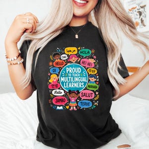 Peut inclure: Un t-shirt noir avec un motif coloré portant le texte PROUD TO TEACH MULTILINGUAL LEARNERS. Le design présente des illustrations d'enfants divers et des bulles de dialogue contenant diverses salutations internationales en plusieurs langues.
