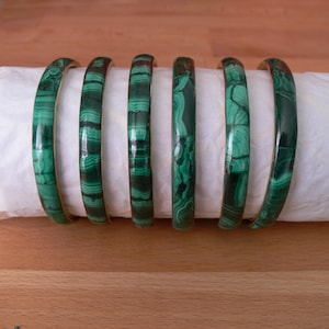 Può includere: Sette braccialetti di malachite verde con finitura lucida. I braccialetti presentano un motivo unico di vortici e linee verde scuro e verde chiaro. I braccialetti sono esposti su una superficie bianca, con sfondo in legno.