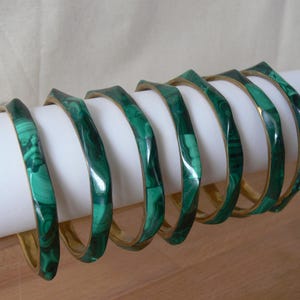 May include: A collection of seven green malachite bracelets with gold-colored metal accents. The bracelets are displayed on a white cylindrical object, showcasing their unique, square-like shape and swirling patterns.