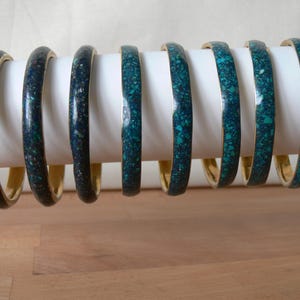 Può includere: Otto braccialetti con interno color oro ed esterno screziato turchese e blu scuro. I braccialetti sono esposti su un cilindro bianco, su sfondo neutro.