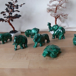 Peut inclure: Une collection de figurines d'animaux en malachite, dont des éléphants, des lions, des rhinocéros et une tortue, est exposée sur une surface en bois. Deux arbres décoratifs en fil de fer avec des perles noires et transparentes sont en arrière-plan. Les figurines sont d'un vert vif.