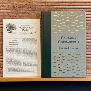 May include: A vintage hardcover book with a gold and gray cover featuring the title "Captains Courageous" by Rudyard Kipling. The book is set against a cork background.