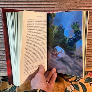 May include: An open book with a red cover and pages featuring text and a colorful illustration. The illustration depicts a large green figure looming over a smaller figure. The text on the page is from Stephen King.