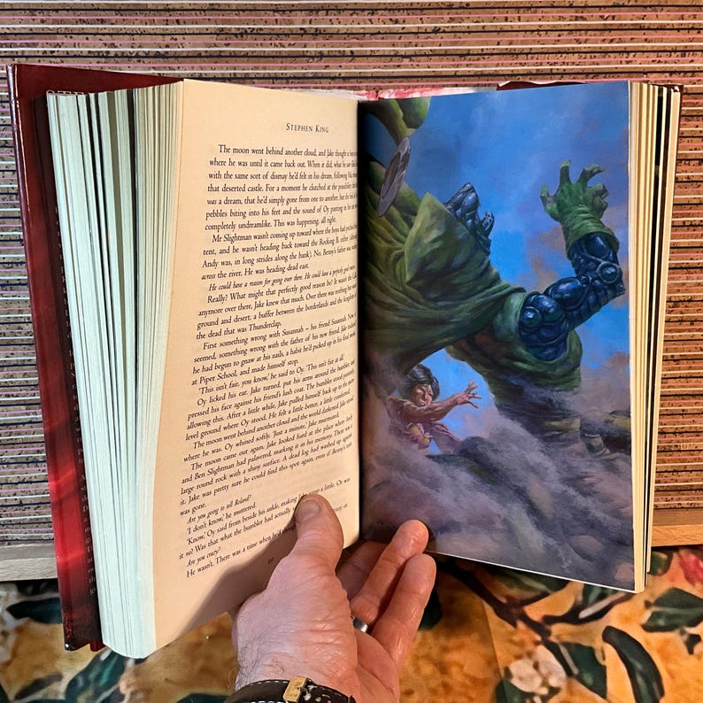 May include: An open book with a red cover and pages featuring text and a colorful illustration. The illustration depicts a large green figure looming over a smaller figure. The text on the page is from Stephen King.