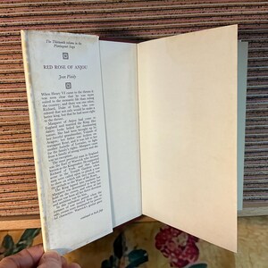 Puede incluir: Un libro de tapa dura abierto, titulado "RED ROSE OF ANJOU" de Jean Plaidy. La portada es blanca con texto negro y lomo rojo. El texto es visible en las p&aacute;ginas abiertas. Forma parte de "The Thirteenth volume in the Plantagenet Saga".