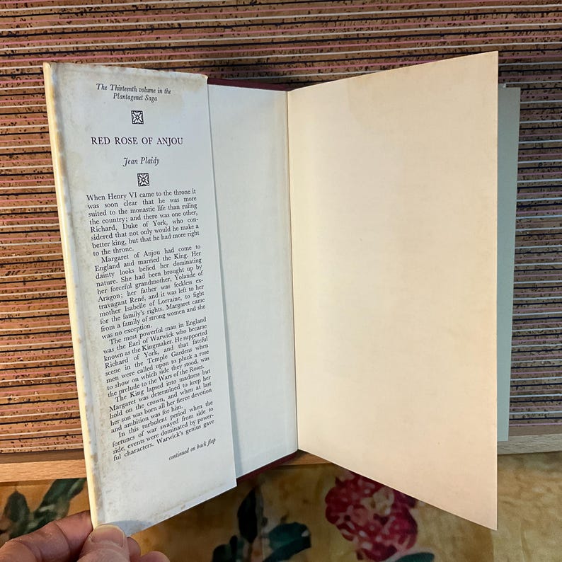 Puede incluir: Un libro de tapa dura abierto, titulado "RED ROSE OF ANJOU" de Jean Plaidy. La portada es blanca con texto negro y lomo rojo. El texto es visible en las p&aacute;ginas abiertas. Forma parte de "The Thirteenth volume in the Plantagenet Saga".