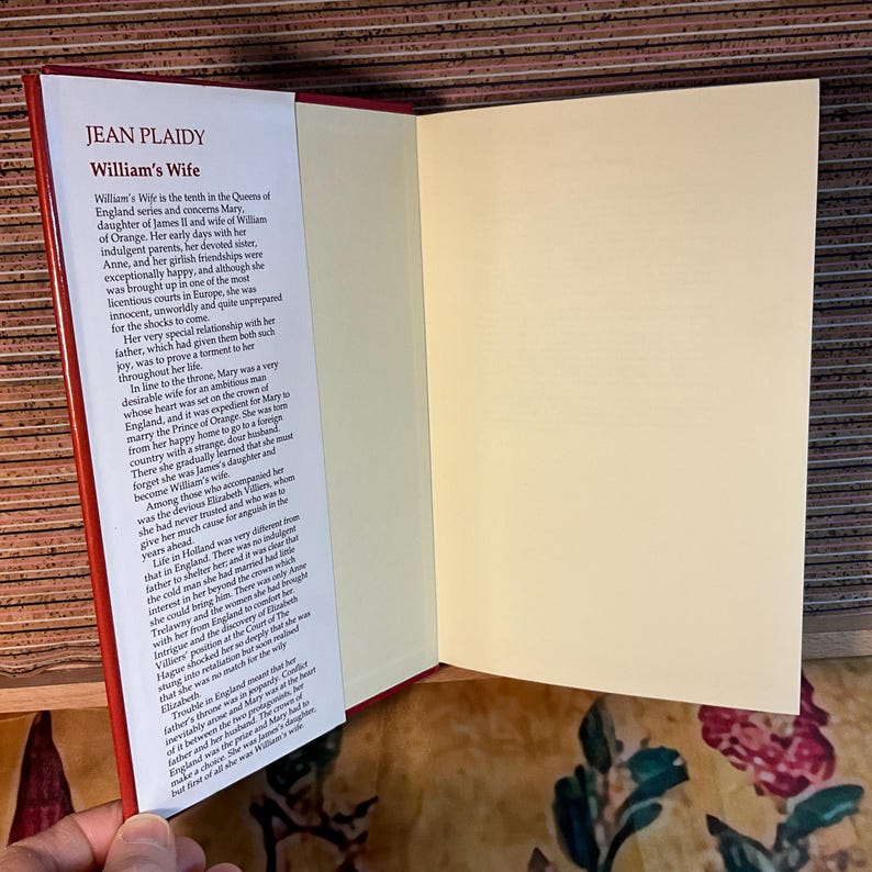 Puede incluir: Un libro abierto titulado "William's Wife" de Jean Plaidy. El libro tiene una cubierta roja y p&aacute;ginas blancas con texto negro. El texto describe la vida de Mar&iacute;a, hija de Jacobo II y esposa de Guillermo de Orange.