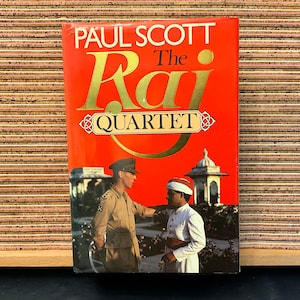 May include: A red book cover with gold lettering that reads "Paul Scott The Raj Quartet". The cover features a photo of two men in period clothing, one in a military uniform and the other in a traditional Indian outfit.