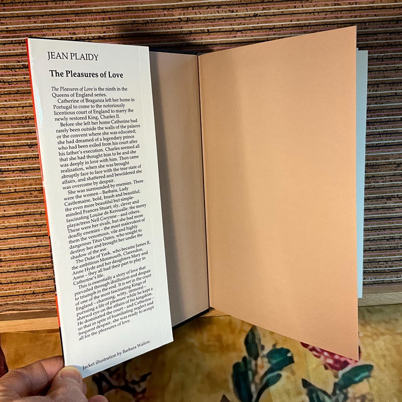 Puede incluir: Un libro abierto titulado "The Pleasures of Love" de Jean Plaidy, de la serie Queens of England. La portada muestra texto y una p&aacute;gina color melocot&oacute;n. El libro revela m&aacute;s texto y un fondo con motivos florales.