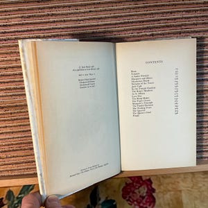Puede incluir: Un libro abierto con p&aacute;ginas color crema. La p&aacute;gina derecha muestra una tabla de contenidos con t&iacute;tulos como "Rene" y "Yolande". La p&aacute;gina izquierda tiene texto sobre la publicaci&oacute;n del libro. El libro est&aacute; sobre una superficie de madera.