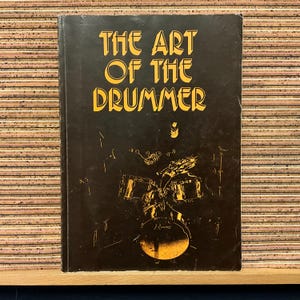 May include: A vintage book titled "THE ART OF THE DRUMMER" in gold lettering on a black background. The cover features a gold illustration of a drum set. The book is rectangular and appears to be in good condition.