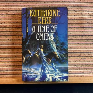 May include: A paperback book cover with a blue background featuring a woman riding a horse through a forest. The title is "A Time of Omens" by Katharine Kerr. The book is part of the "Westlands Cycle" and is book two of the series.
