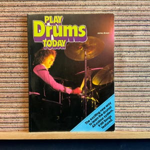 May include: A book cover with the title "Play Drums Today" in purple and yellow lettering. The cover features a photo of a person playing a drum set. The text "Ashley Brown" is in the upper right corner. The text "The exciting new way to learn the drums! All-colour system. 9 Hit Songs featured." is in the lower right corner.