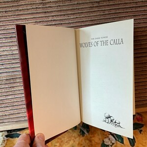 May include: Open book with a red spine and white pages. The text "THE DARK TOWER WOLVES OF THE CALLA" is printed on the right page. A small illustration of several round objects is at the bottom of the right page.