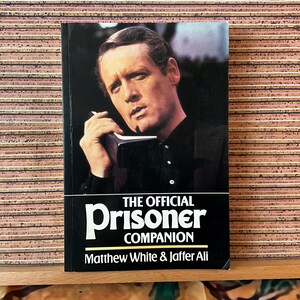 May include: A paperback book titled The Official Prisoner Companion by Matthew White and Jaffer Ali. The cover features a photograph of a man in a black shirt holding a black telephone receiver to his ear against a dark background.