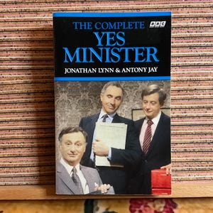 Könnte beinhalten: Ein gebundenes Buch mit dem Titel "The Complete Yes Minister" von Jonathan Lynn & Antony Jay. Das Cover zeigt einen blauen und schwarzen Hintergrund mit dem BBC-Logo und Bildern von drei Männern in Anzügen.