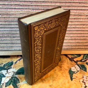 May include: A vintage book with a brown leather cover and gold decorative detailing. The cover has an ornate gold border and a central panel. The pages are aged, and a cream ribbon bookmark is visible.
