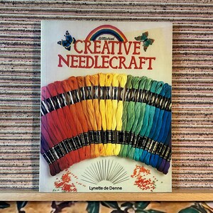 May include: A book titled "Creative Needlecraft" with a rainbow and butterfly design. The book displays a fan of colorful embroidery floss in shades of red, orange, yellow, green, blue, and purple. The author's name, Lynette de Denne, is also visible.