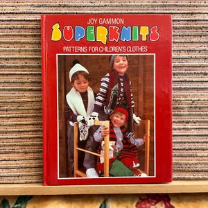 May include: A red book cover with the title "Superknits Patterns for Children's Clothes" by Joy Gammon. The image shows three children in knitted winter wear, including scarves and hats. The children are smiling and appear to be happy.