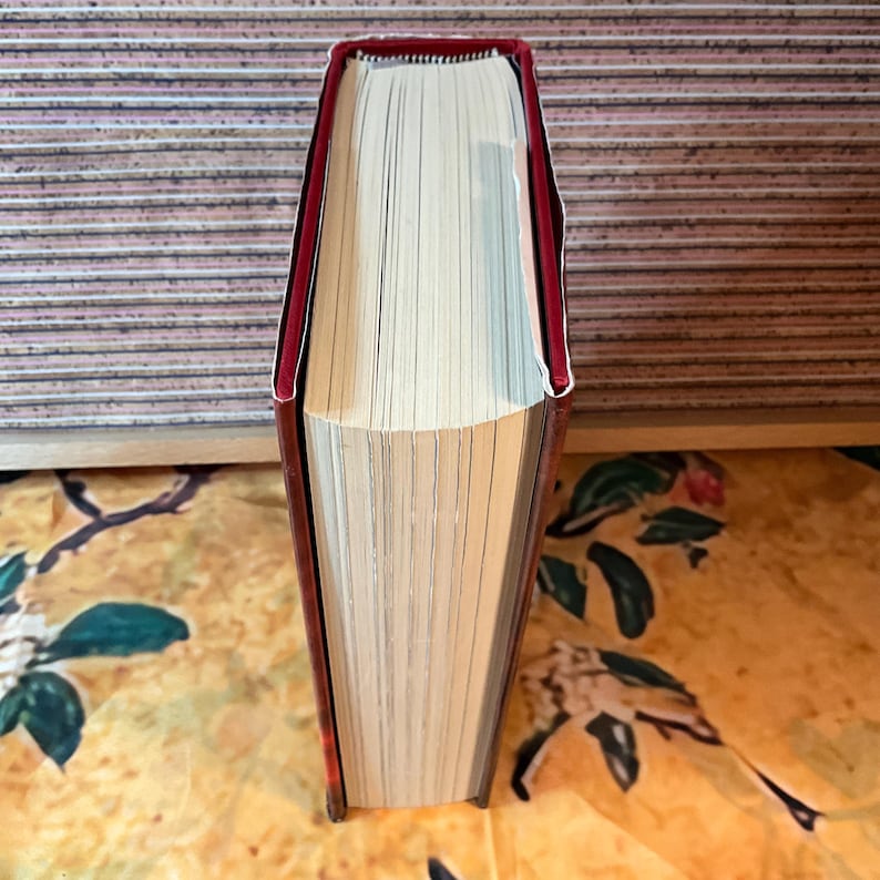 May include: A hardcover book with a red spine and edges. The pages are cream-colored and the book is closed. The book is standing upright, showing the spine and the top edge of the pages.
