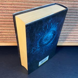 May include: A hardcover book with a blue and black cover featuring a stylized eye with a circular design. The book's spine is a light brown color.