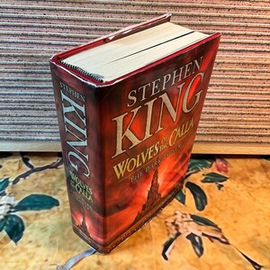 May include: A hardcover book titled "Wolves of the Calla" by Stephen King. The cover is predominantly dark red with the title in large, bold, orange letters. The book has a bookmark and is set against a patterned background.